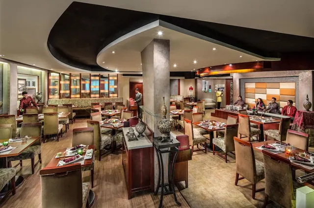 A restaurant dining room with wooden tables and upholstered chairs, a central pillar, and guests seated at a counter in the background.