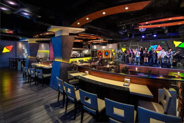 A lounge with a curved bar counter and upholstered chairs, with a live band performing on a raised stage beside colourful wall graphics.