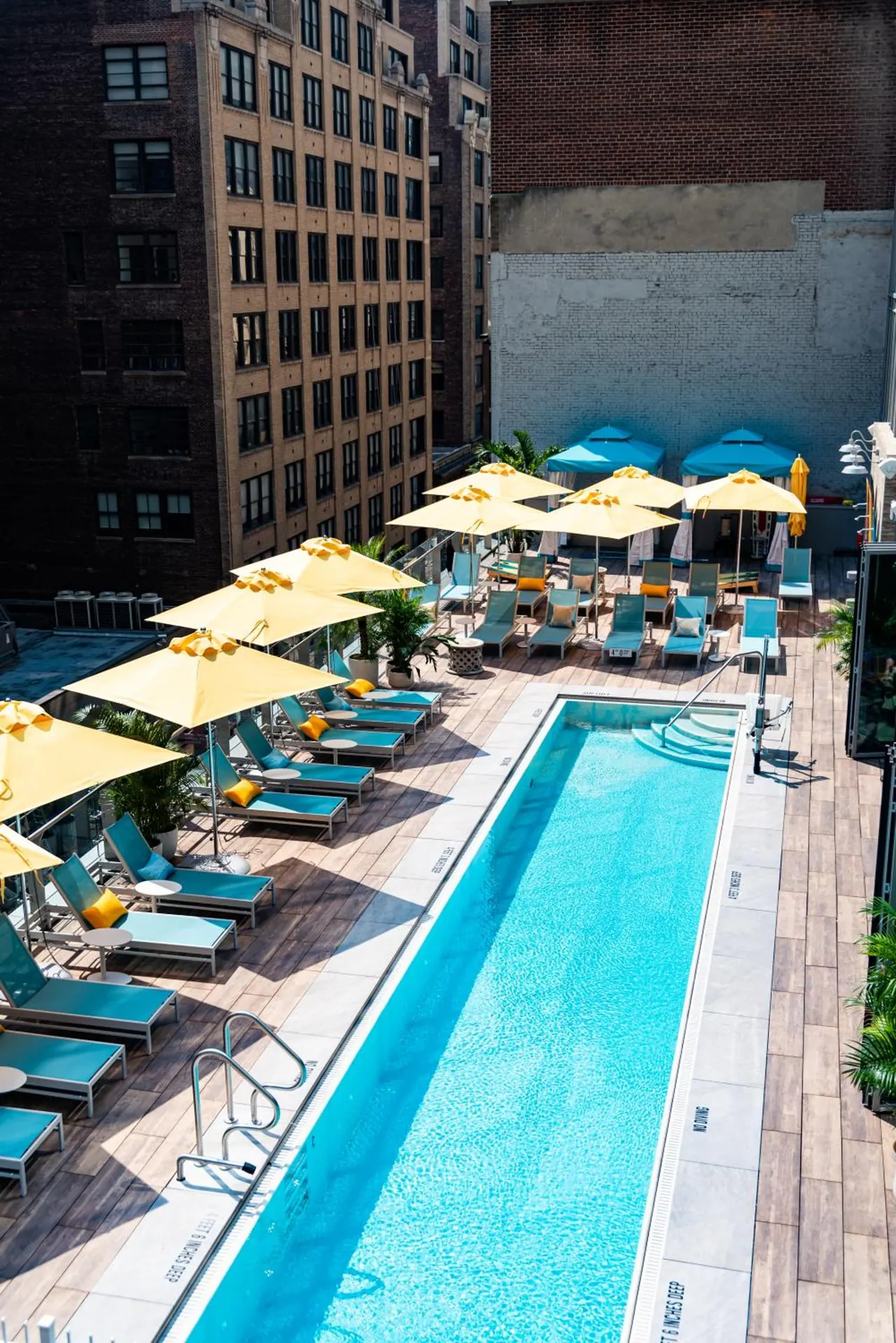 A rooftop swimming pool with yellow umbrellas and teal loungers, surrounded by brick buildings.