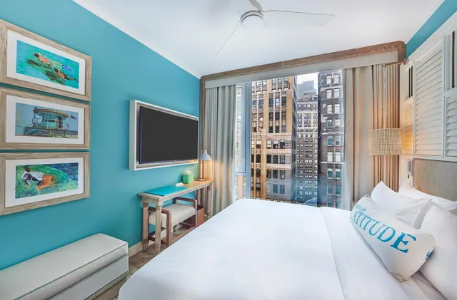 A bedroom with a white bed, a wall-mounted television, a desk with a chair, and a large window looking onto tall city buildings.