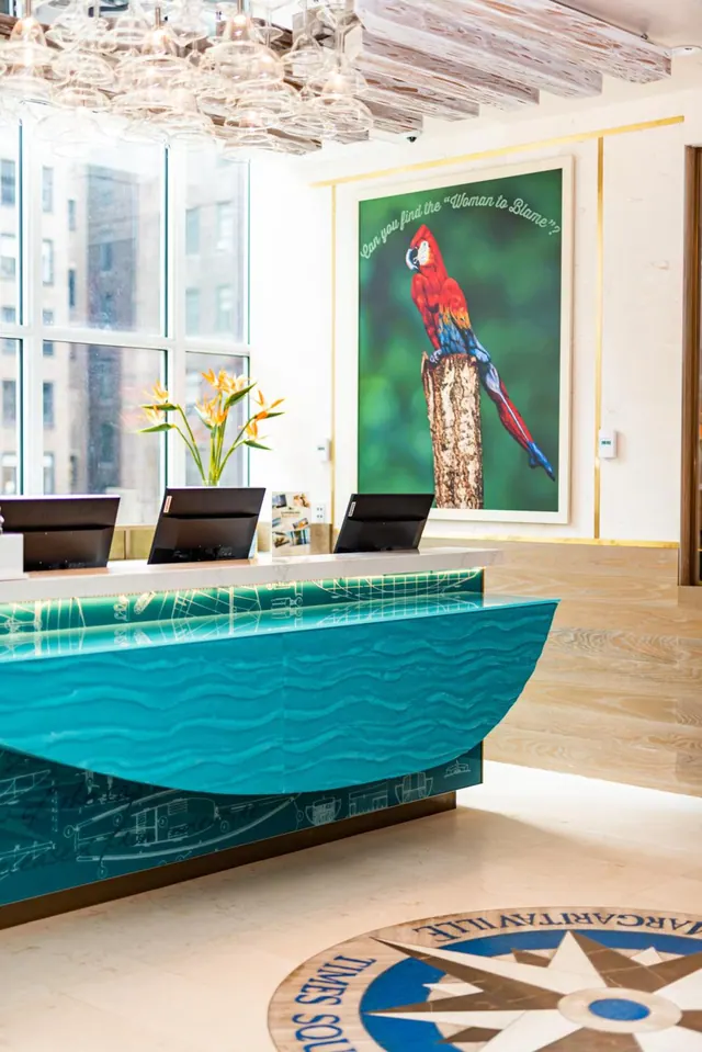 A hotel lobby reception desk with a teal wave-textured front and two computer monitors, with a large parrot artwork on the wall and a compass rose design in the floor.