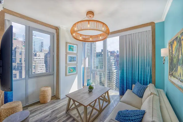 A bright living area with a light sofa, a wooden coffee table, a wall-mounted television and large windows looking out over city buildings.