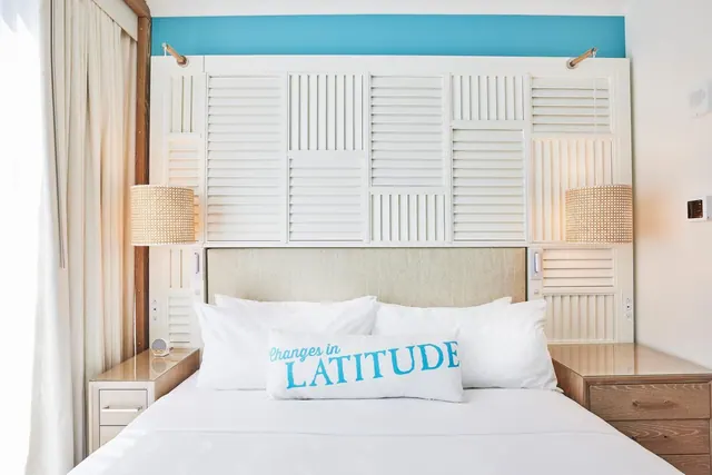 A made bed with white linens and a turquoise-lettered pillow sits in front of a shutter-panelled wall with wicker pendant lamps on either side.