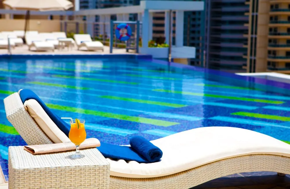 A cocktail, folded blue towels and a wicker side table sit on a cushioned lounger beside a blue rooftop pool, with high-rise buildings behind.