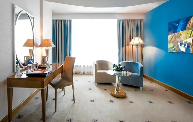 A hotel sitting area with a wooden desk, mirror, two table lamps, two armchairs, a round glass table and a bright blue accent wall.