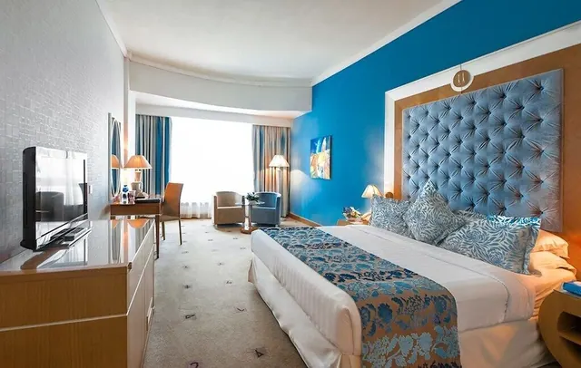 A hotel bedroom with a king bed under a tufted blue headboard, a television on a cabinet, a desk with chair, and two armchairs by a bright window.