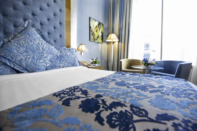 A bedroom with a made bed under a blue floral throw, two armchairs beside a small round table, table lamps and a bright window with curtains.