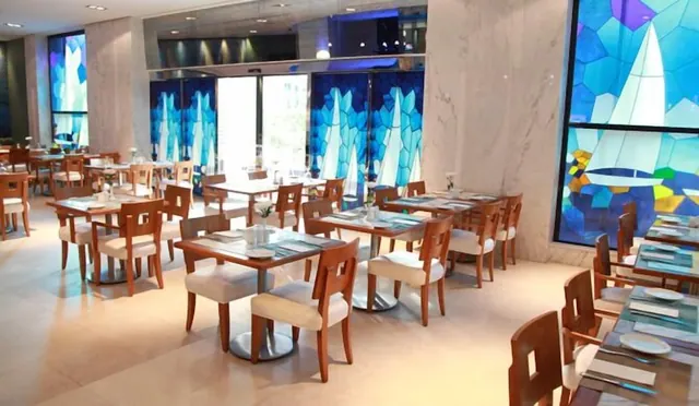 A restaurant dining room with wooden chairs and set tables beneath large blue stained-glass panels showing sailboats.