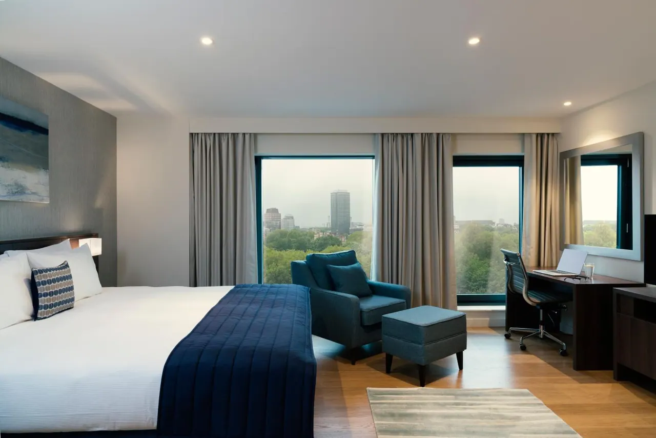 A hotel bedroom with a king bed, a blue armchair and ottoman, a work desk, and windows looking out over trees and tall buildings.