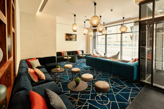 A lounge with dark teal sofas, low round tables, a patterned blue carpet and pendant lights, with large windows looking onto a street.