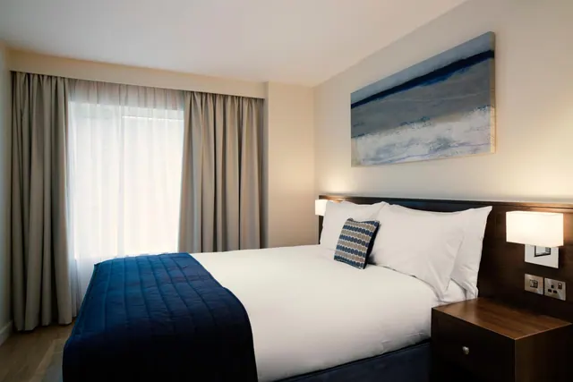A made double bed with a navy throw and patterned cushions sits beside a dark wood bedside table, below a blue-toned painting, with a sheer-curtained window to the left.