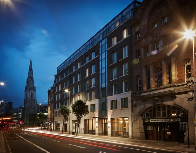 A modern dark brick hotel building stands beside a Victorian stone building on a street at dusk, with a church spire in the distance and light trails on the road.