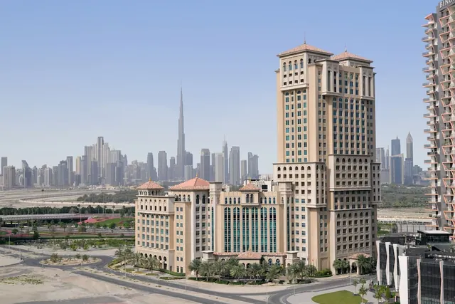 Exterior of a beige hotel complex with palm trees and roadway in front, with a city skyline including the Burj Khalifa in the background under a clear sky.