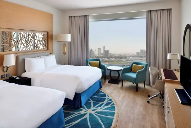 A hotel bedroom with two large beds, armchairs beside a window overlooking a city skyline, and a desk with a wall-mounted television.