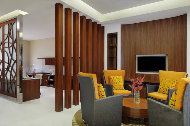 A living area with four yellow-cushioned armchairs around a round wooden coffee table holding a vase of pink flowers, with a wall-mounted television and a desk visible behind a wooden slatted divider.