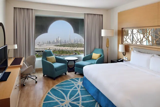 A hotel bedroom with a king bed, teal armchairs, a work desk and an arched window looking out over the city skyline and Burj Khalifa.