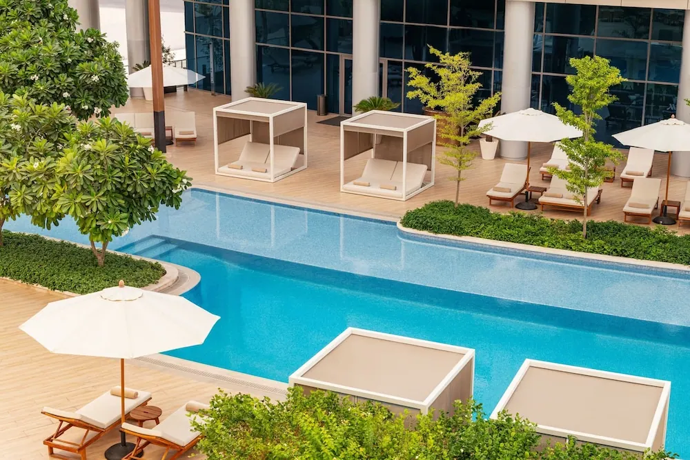 An outdoor pool with blue water sits beside a deck with white canopied daybeds, sun loungers under parasols, and green planting.