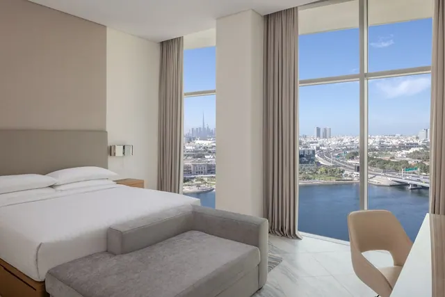A bedroom with a king bed, a grey bench at the foot of the bed, a desk and chair, and floor-to-ceiling windows overlooking a waterway and a city skyline with a distant tower.