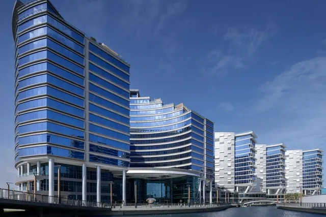 Exterior view of several glass-fronted towers beside a canal, with a footbridge and walkway in the foreground under a clear blue sky.
