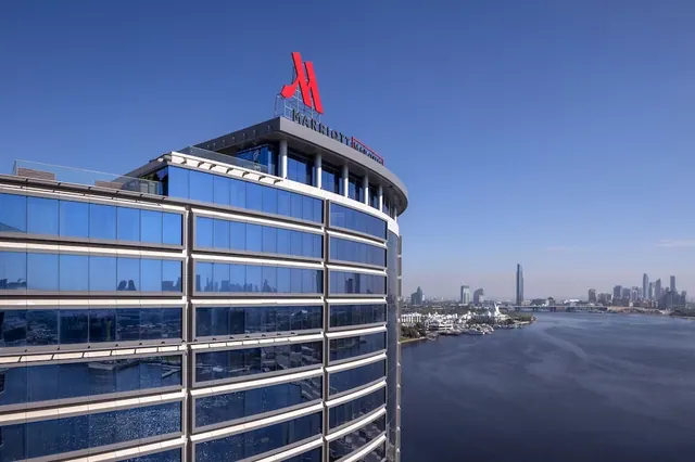 Exterior of a glass-fronted hotel tower with a red Marriott logo on its roof, overlooking a waterway and city skyline under a clear blue sky.