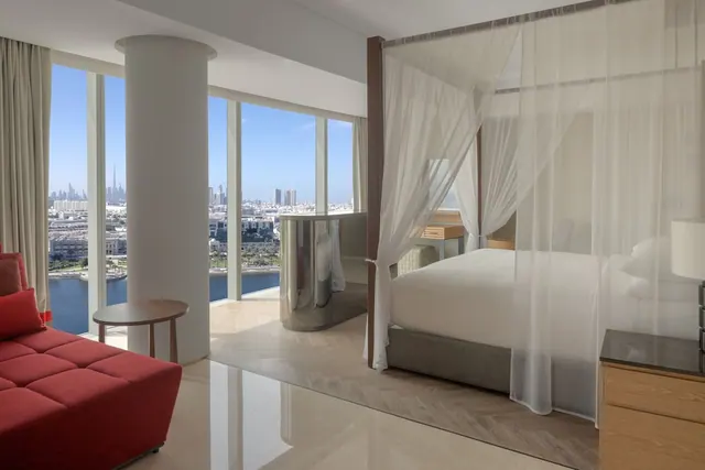 A bedroom with a canopy bed behind sheer drapes, a red sofa, and floor-to-ceiling windows overlooking a city skyline and a waterway.