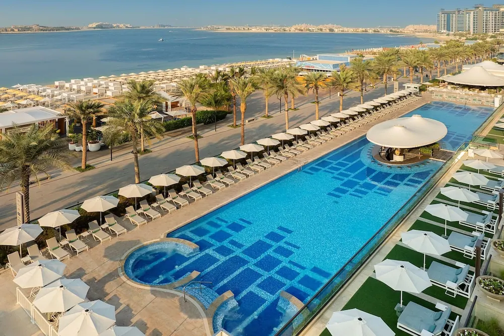 Aerial view of a large outdoor pool with a round hot tub, rows of sun loungers and white umbrellas, with the sea and beach beyond.