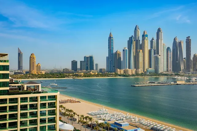 View from an elevated building over a sandy beach with white umbrellas and sun loungers, the sea, a pier, and a high-rise skyline in the distance.