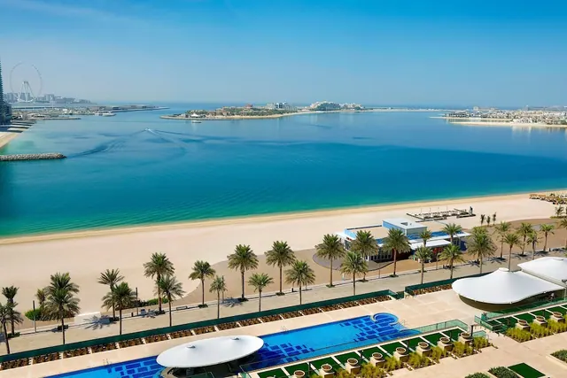 Elevated view over a pool, palm-lined sandy beach and calm turquoise sea toward the Ain Dubai ferris wheel at left.