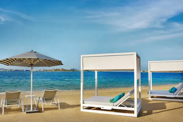 White cabana daybeds with teal cushions and a striped umbrella stand on a sandy beach facing the sea.