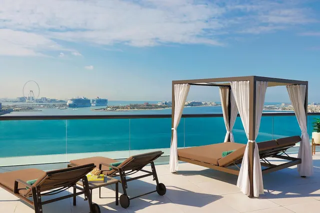 A covered daybed and two sun loungers sit on a tiled terrace overlooking the sea, with cruise ships and the Ain Dubai observation wheel in the distance.