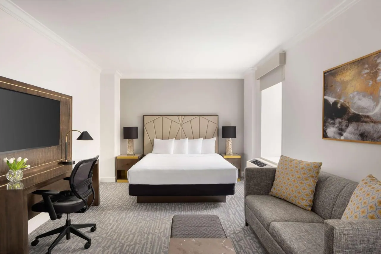 A hotel bedroom with a king bed, a wall-mounted television, a desk with a chair, and a grey sofa with patterned cushions.