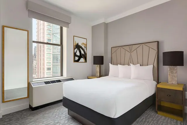 A king bed with white bedding and a tufted headboard sits beside a window that looks out onto a high-rise building, with a wall unit below the window.
