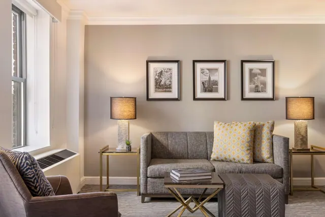 A living area with a grey loveseat, a brown armchair, two table lamps and three framed black and white prints of New York buildings on the wall.