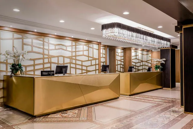 A hotel lobby reception area with two gold-coloured front desks, a linear crystal chandelier, white orchid arrangements and a Hilton Curio Collection sign on the desk.