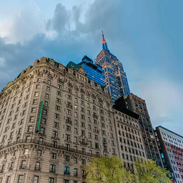 Low-angle view of a historic stone building with ornate cornices on a city street, with the Empire State Building rising behind it under a cloudy blue sky.