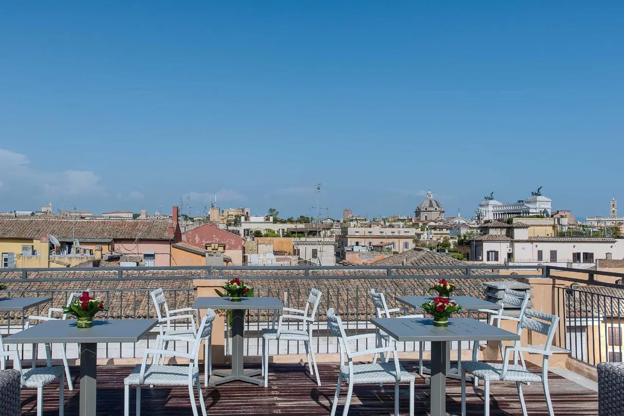 A rooftop terrace with grey tables, white chairs and flower arrangements overlooks Rome's rooftops and the white Vittoriano monument under a clear blue sky.