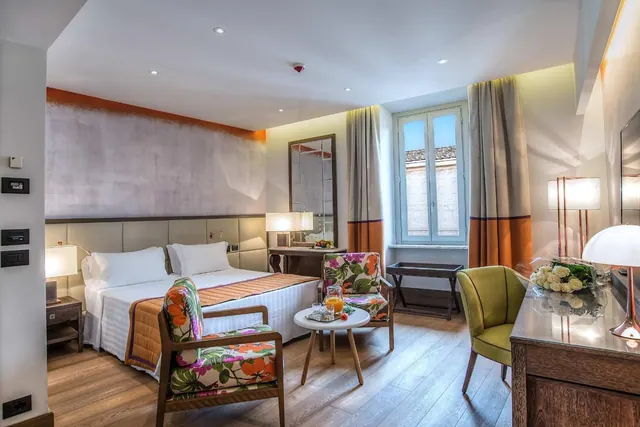 A double bed with an orange throw sits beside two floral armchairs, a small round table with a glass of juice, and a window looking onto a neighbouring building.