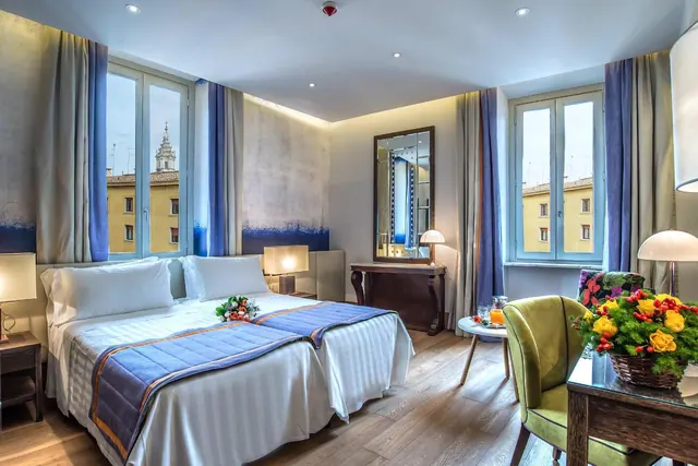 A hotel bedroom with two beds dressed in white linen and blue runners, tall windows overlooking a yellow building with a church tower, and a green armchair beside a desk with a flower arrangement.