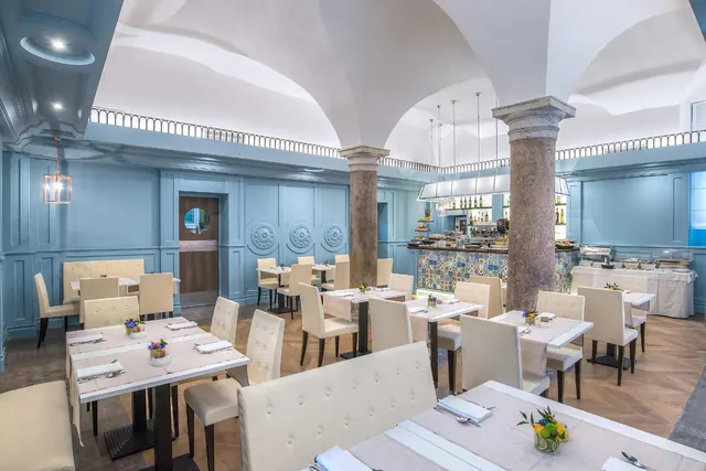 A restaurant dining room with blue walls, a vaulted ceiling, stone columns, set tables with cream tufted chairs, and a buffet counter at the back.