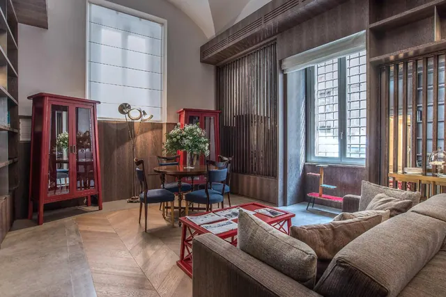 A living area with a grey sofa, a red coffee table, red display cabinets, a round dining table with blue upholstered chairs, and a window with a grille.