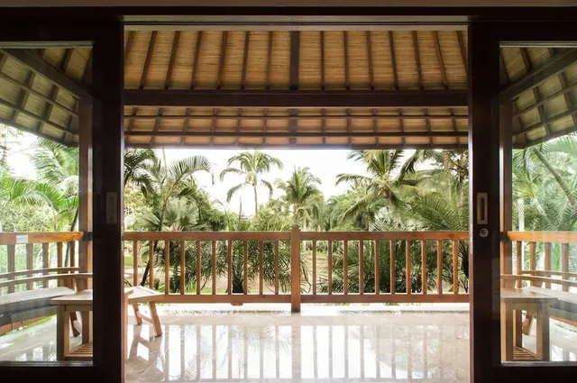 View through open wooden doors from inside a room onto a tiled terrace with a wooden railing and palm trees beyond.