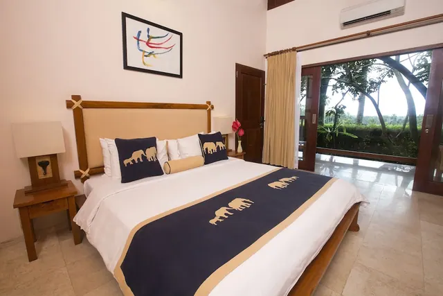 A bedroom with a king bed in navy and cream linen, wooden bedside tables, an air conditioner on the wall, and glass doors opening onto trees and greenery.