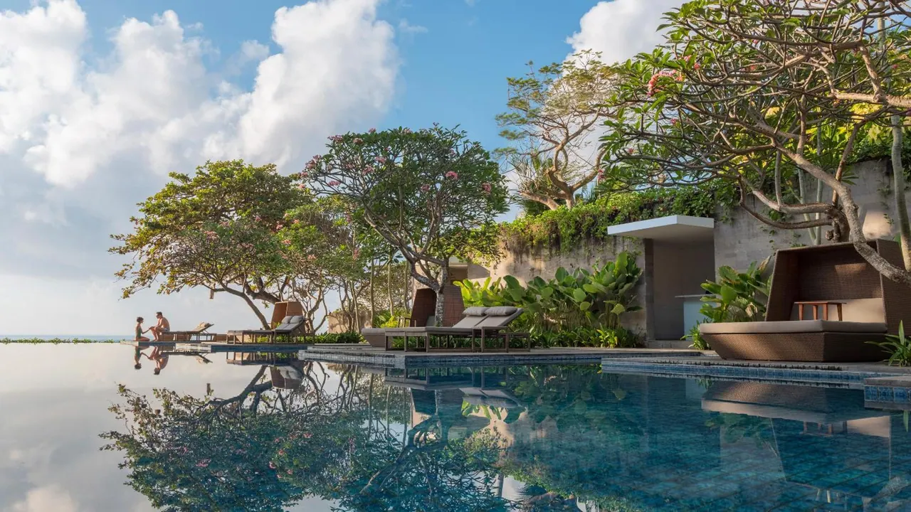 An infinity-edge pool with sun loungers and a daybed beside it, frangipani trees, and the sea horizon visible beyond, with two people seated at the far edge.
