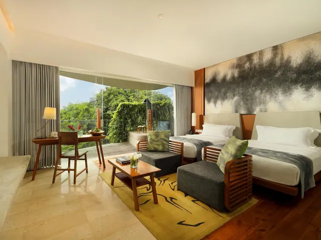 A hotel bedroom with two beds, armchairs, a desk and a glass wall opening onto a balcony with a leafy garden view.
