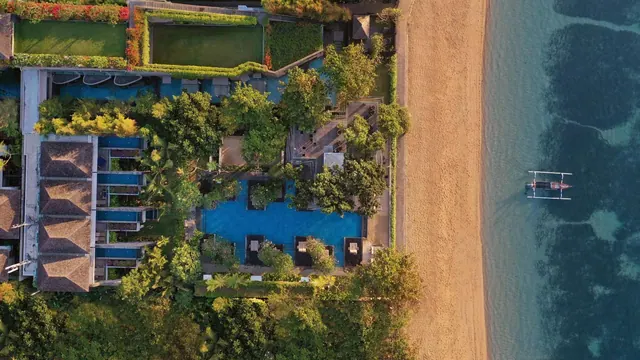 Aerial view of a resort with a central swimming pool, small pools beside thatched-roof villas, a sandy beach and an outrigger boat on the sea.