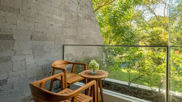 A small balcony with two wooden chairs and a round wooden table holding white flowers, beside a stone-clad wall and a glass railing overlooking a garden of trees and lawn.