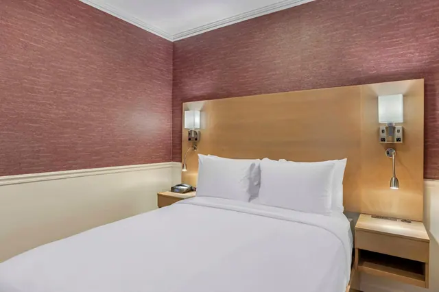 A made double bed with white linens and a wooden headboard, with wall-mounted reading lights and a telephone on the bedside tables.