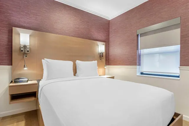 A bedroom with a made double bed with white linens, wall-mounted reading lights, a nightstand with a clock radio, and a window with a roller blind partly lowered.