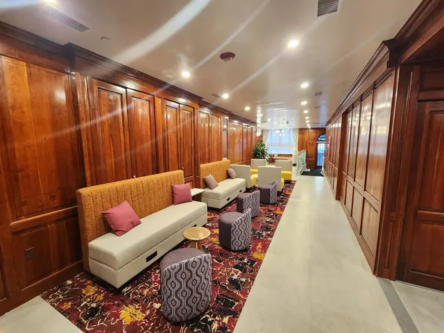 A wood-paneled hallway with cushioned banquette seating, round ottomans and a patterned red rug, leading toward a window at the far end.