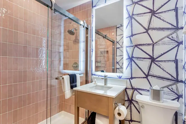 A bathroom with a glass-enclosed walk-in shower lined with pink tiles, a wood-base vanity under a lit mirror, and a toilet beside geometric patterned wallpaper.